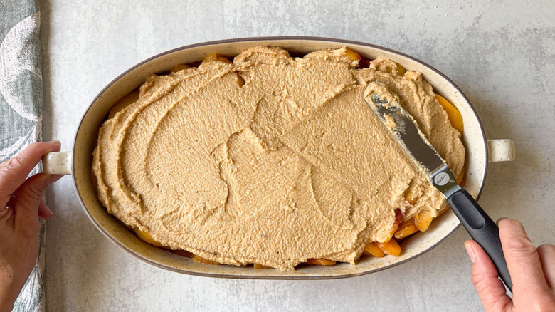 Spreading gluten-free cobbler dough over sliced peaches in oval baking dish