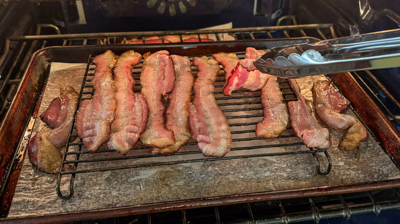 Bacon on sheet pan with baker's rack in oven with tongs turning the bacon slices