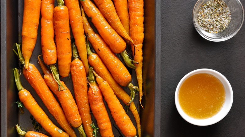 A pan of whole, honey-glazed carrots on a black countertop, a ramekin of salt and pepper and one of honey, to the side