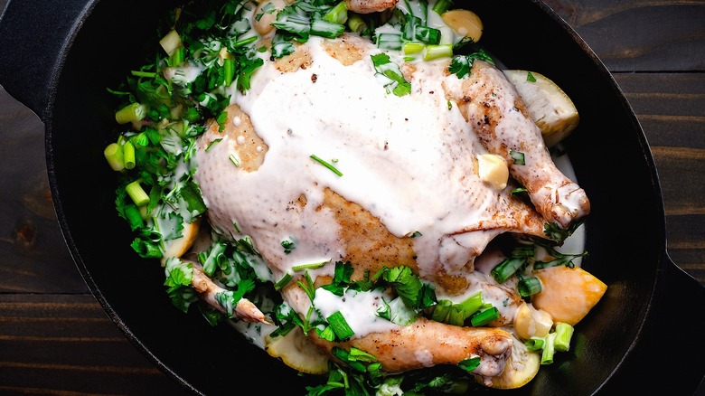 Whole chicken in black pot covered in fresh herbs, green onions, and milk