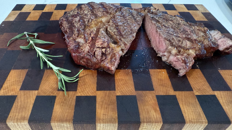 A cooked ribeye steak on a butcher block with a sprig of rosemary